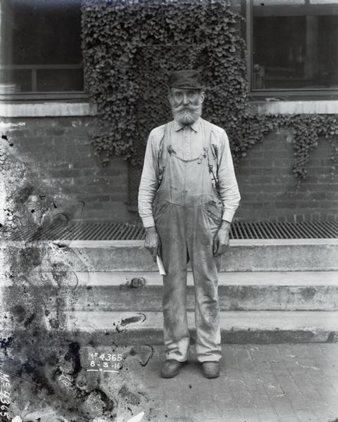 A man in worn overalls and a dark cap is standing outside a brick building. The man is most likely a factory worker at International Harvester's McCormick Works.
