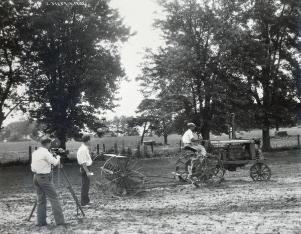 A man is operating a Farmall tractor and grain drill in a field. One man is looking on, and another man is filming the process with a movie camera.