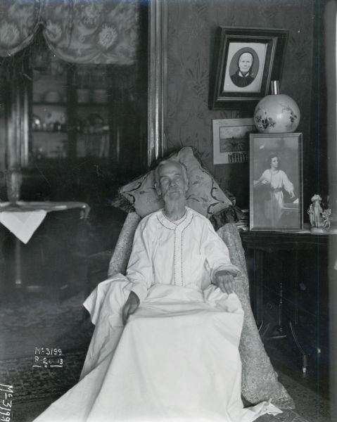 Portrait of a seated man wearing a white dressing gown, covered with a white blanket. He is resting his head against a pillow in what appears to be a domestic setting, with portraits, a lamp, and a small figurine behind him. The man may be an employee of International Harvester's McCormick Works (factory).