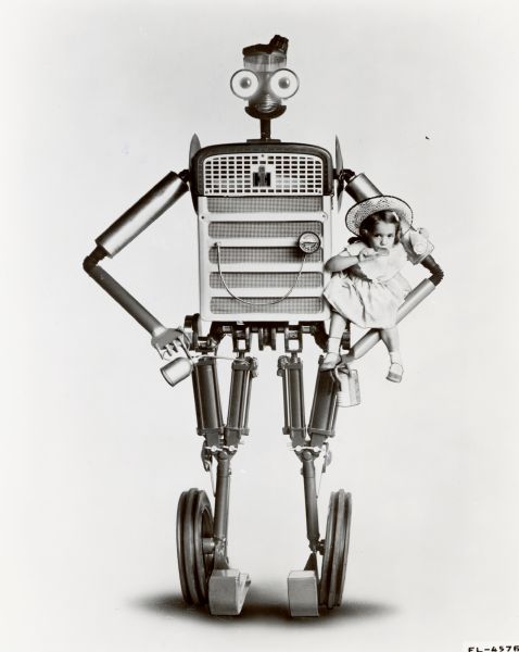 A young girl eating a lollipop is perched in the crook of the arm of "Tracto," a robot made of tractor parts. Tracto was part of International Harvester exhibits at state fairs. From the original press release: "the eight-foot 'talking' robot [is] assembled from 227 farm tractor and implement parts. Robot is equipped with two-way communication system and contains motor gear reducer for oscillating head and lifting right arm. Electrically powered, eyes light up through transformer installed in robot's stomach." According to another press release, Tracto was voiced by "district office personnel hidden from the view of passers-by, but advantageously located to spot interested viewers and provoke them into conversations."