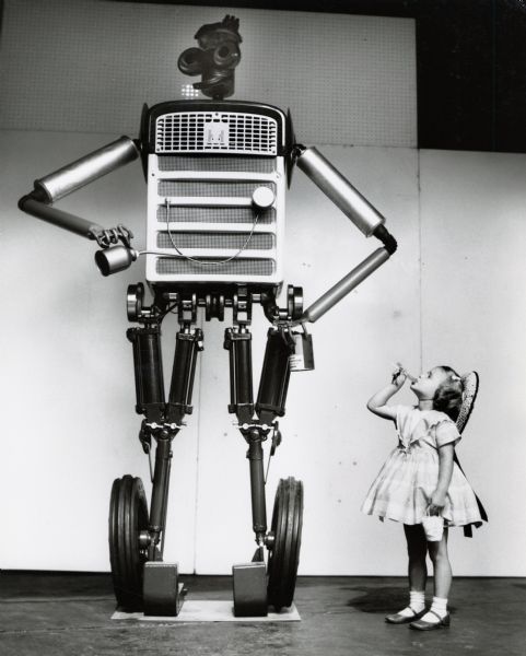 A young girl with a lollipop looks up at "Tracto," a robot made of tractor parts for International Harvester's exhibits at state fairs. 
From the original press release: "the eight-foot 'talking' robot [is] assembled from 227 farm tractor and implement parts. Robot is equipped with two-way communication system and contains motor gear reducer for oscillating head and lifting right arm. Electrically powered, eyes light up through transformer installed in robot's stomach." According to another press release, "Tracto" was voiced by "district office personnel hidden from the view of passers-by, but advantageously located to spot interested viewers and provoke them into conversations."
