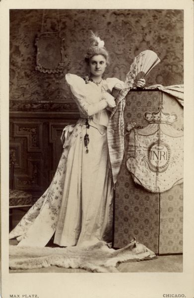Full-length carte-de-visite of Anita McCormick Blaine (1866-1954) wearing a formal gown and carrying a fan. Anita's hair is powdered, and she stands by a screen with what seems to be a royal crest, that reads: "Inri", and with an animal-skin rug at her feet. It is possible that she is in costume as Marie Antoinette.