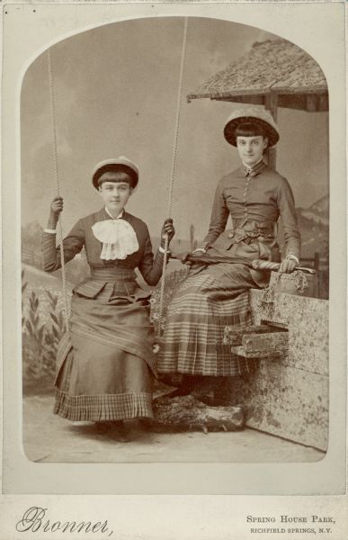 Carte-de-visite of Anita McCormick Blaine (1866-1954) and an unidentified young woman. Anita sits on a "well" and the friend sits on a swing in front of a painted backdrop of a country scene. The young women both wear long skirts, hats, and gloves.