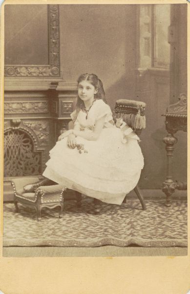 Studio portrait of a young Mary Virginia McCormick (1861-1941) in front of a painted backdrop. She is wearing a white dress with a striped sash, and sitting on a child-size chair with her feet on a small footstool.