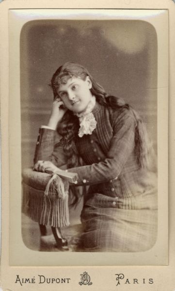 Carte-de-visite of a three-quarter length studio portrait of Anita McCormick Blaine (1866-1954) as a young woman sitting in a chair. She is posing leaning her head on her hand, and is holding a fan in the other hand on the arm of a chair. She is wearing a plaid dress with a lace collar.