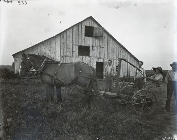 A boy wearing a large floppy hat is sitting on a sickle-bar mower hitched to a team of horses. An older man, with a beard and a straw hat, and two younger men are standing beside the mower, in front of a large barn. An inscription describes it as a "farm family posed in front of old barn." The two younger men and the boy are wearing gingham shirts. All four men wear soft collar shirts and suspenders.