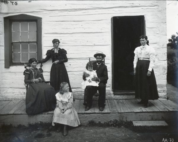 A man, three women and two young girls posing on the porch of a whitewashed log house. The man is wearing a suit and straw boater hat, the women are wearing long dresses.