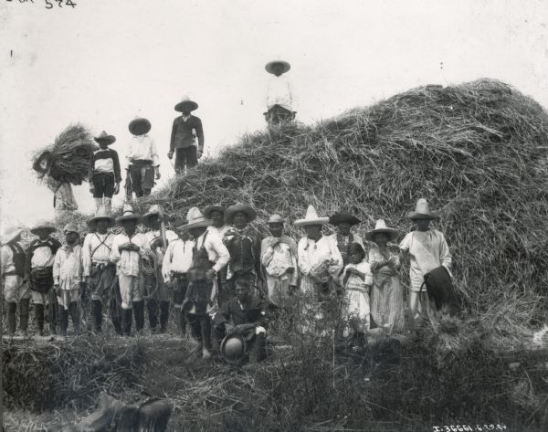 Group of Mexican workers posing in front of a large stack of hay. Most of the people are wearing hats, many of them sombreros, and worn clothing. Several people are carrying hand tools, including ropes, a sickle, and a wooden rake.