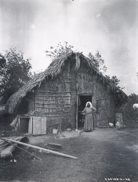 Woman standing in the open doorway of a house that appears to be constructed of woven reeds attached to columns.  The roof of the hut is thatched.  A variety of debris and several chickens are in front of the house.