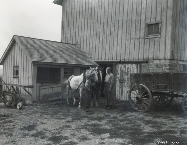 Boy speaking to man while holding a team of horses near a large barn. A McCormick-Deering Weber wagon, manufactured by International Harvester, and a plow, are both beside the barn.