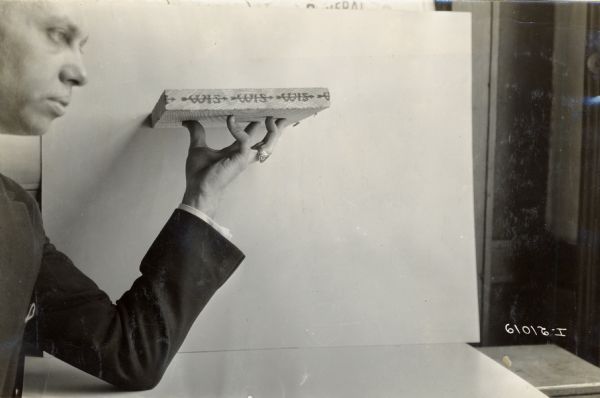 A man holds a wooden board reading "WIS" against a white backdrop. The "WIS" may stand for Wisconsin Lumber Company, a subsidiary of International Harvester. It's possible that board was a surrogate for an object that would later be added by an artist, and would appear in an advertisement.