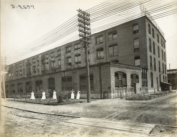 View across railroad tracks towards a group of five women in white aprons standing at the side of a factory building, possibly Deering Twine Mill. Other workers are watching the photographer through open windows on the side of the factory building.