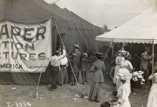 A small crowd outside a large tent that adverstises "Reaper Motion Pictures." The tent is likely part of an International Harvester exhibit at a fair or agricultural equipment demonstration. Children, including two in baby carriages, and women in large hats make up the crowd outside of the tent.
