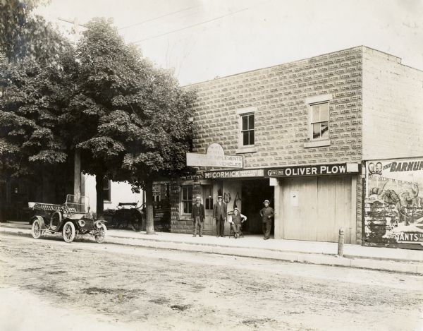 A group of four men are standing outside the Newell McCormick Implements and High Class Vehicles dealership in Ontario, Canada. A motor car is parked in the street outside the store, and an advertisement for the Barnum and Bailey circus is attached to a wall next to the dealership.