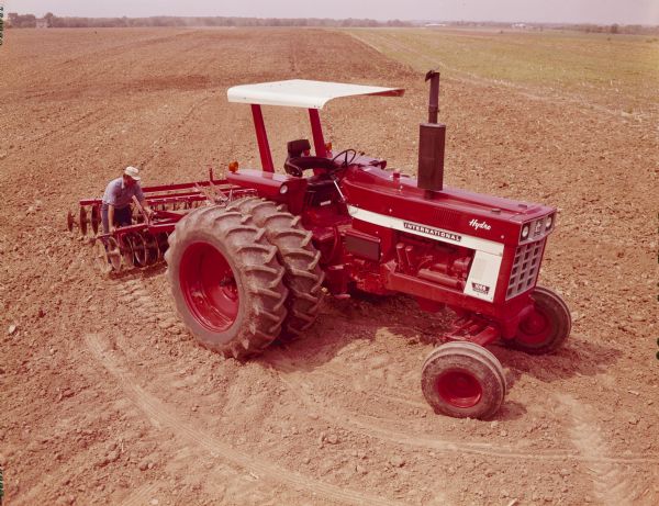 Color photograph of a man standing at the back of an International Hydro 1066 tractor adjusting a 370 Disc, 13 foot and 10 inch Disc Harrow attachment.