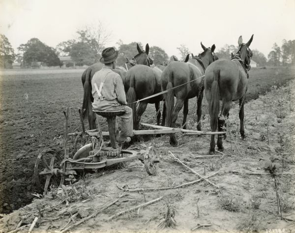 Rear view of a man plowing a field with a Chattanooga reversible(?) plow pulled by four horses.
