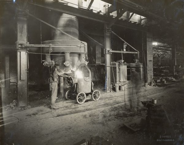 Factory worker in a foundry at the Chattanooga Plow Works fills a cart with hot metal. A blurred image of another worker that passed through the scene while the photograph was being taken is just right of center.