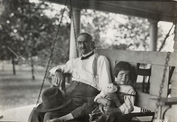 A older man and a young girl holding a doll are seated together on a porch swing for an outdoor portrait.