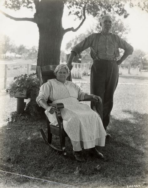 A man stands next to a woman seated in a rocking chair for an outdoor portrait. The man and woman may be employees or former employees of International Harvester.