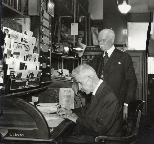 Two men look over papers at a desk surrounded by International Harvester manuals, pamphlets, and a repair list. The men may be International Harvester dealers, or branch house office workers.