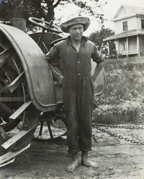 A young boy wearing a hat and coveralls standing barefoot next to a tractor.