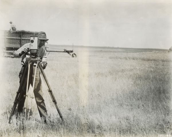 A photographer, likely an employee of International Harvester, and his hand-crank motion picture camera standing in a field in front of man on top of a wagon.