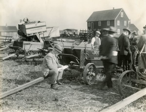 A man examining papers sits on the beam of an farm wagon (probably a John Deere) while others gather near him. A variety of farm machines are in the background. The men may be attending a farm machine demonstration or perhaps an estate sale.