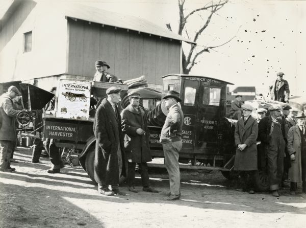 Mr. Porter of the Porter Hardware Co., an International Harvester dealership, stands in front of an International Red Baby truck while other men mill around. The truck appears to be loaded with a feed grinder, along with a manure spreader advertising poster.