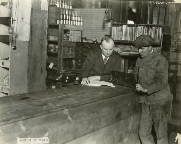 International Harvester dealer W.E. Tomlinson examines what appears to be a machine manual in a repair shop or dealership while another man looks on.