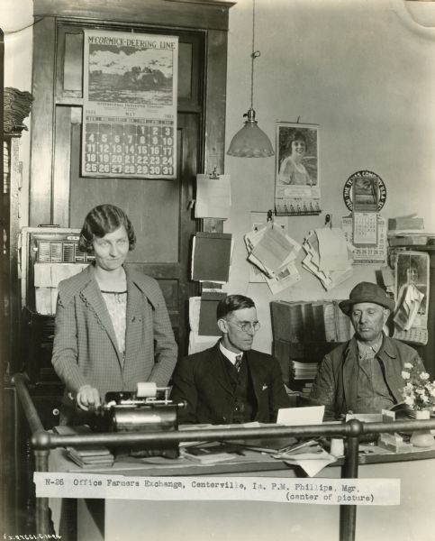P.M. Phillips, manager of the Office Farmers Exchange, sits behind a desk, with a woman typing on one side and a seated man on the other. A McCormick-Deering calendar hangs in the background. The Office Farmers Exchange was likely an International Harvester dealership.