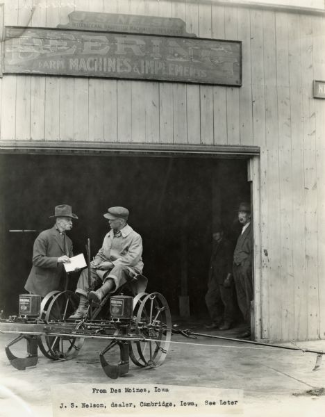 J.S. Nelson, an International Harvester dealer, speaks with a man seated on corn planter(?). Two men smoking pipes watch from the shed in the background.