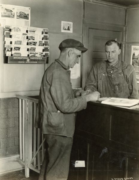 Two men standing at the counter of an International Harvester dealership. Posters and machine pamphlets are hanging in the background.
