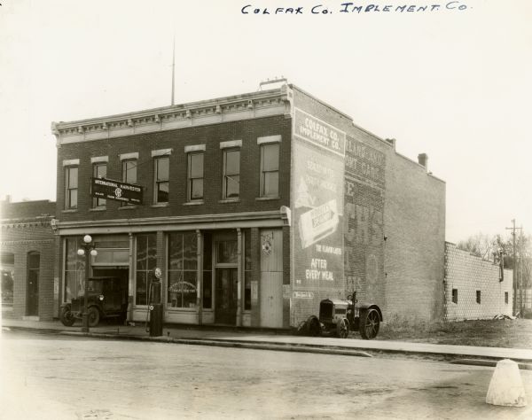 Exterior view of the Colfax Co. Implement Company with a pump for Red Crown gasoline in front of it.  An automobile is emerging from the garage and a tractor sits along the side of the building. The Colfax Co. Implement Company was an International Harvester dealership.