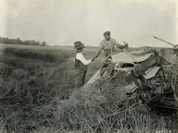 A man seated on a Deering grain binder shakes hands with a man standing and smoking a cigar. The man with the cigar may be an International Harvester dealer.