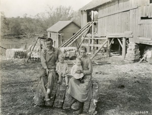 A couple with two children sitting on a bale of cotton in a barnyard posing for a portrait.