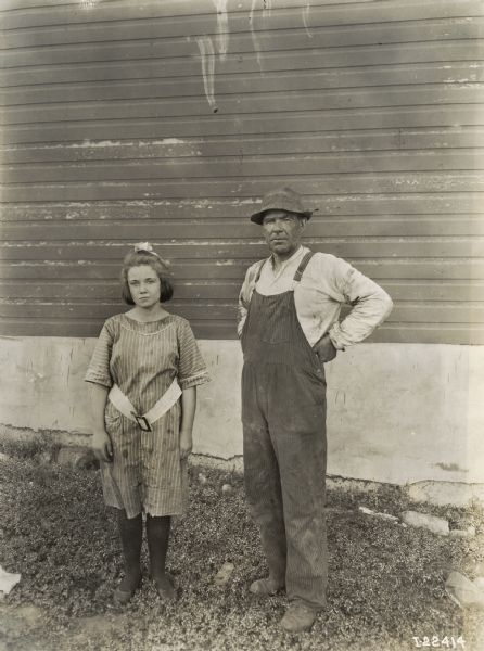 A man in a hat and overalls, and a girl in a dress with a loose belt, stand in front of a building to have their portrait taken. The man is likely a farmer, and the girl may be his daughter.