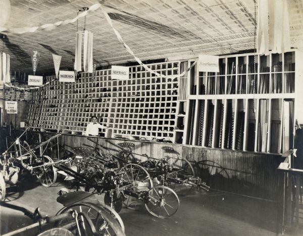 The showroom floor of Dixon Implement Company, an International Harvester dealership. Behind a man standing at the sales counter, and against the far wall, are signs indicating brands sold in the store: Deering, McCormick, Milwuakee and Miscellaneous. The floor is covered with different tractor and implement parts and the ceiling is decorated with paper streamers.
