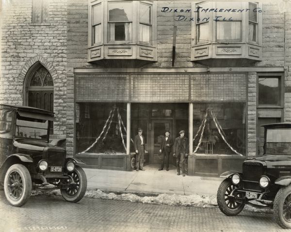 Storefront of the Dixon Implement Company, an International Harvester dealership, with three men standing in the doorway. Two parked trucks frame the storefront. Merchandise is displayed in the two front windows along with paper streamers.