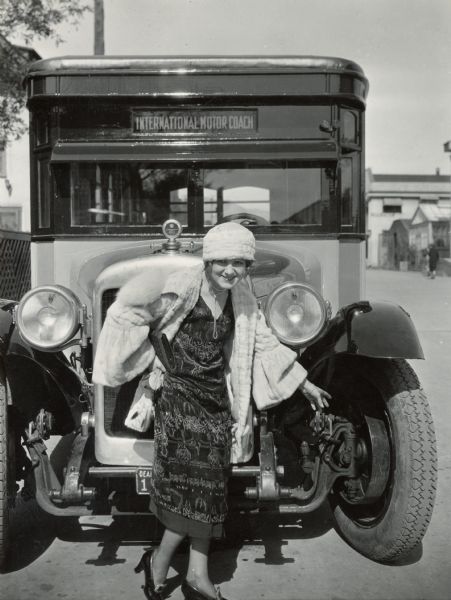 A woman dressed in furs, patterned dress, and high heels is posing in front of an International Harvester motor coach, or bus. She seems to be pointing to the exposed machinery between the front wheel and engine. The bus was built on a model 52 or model 53 chassis.