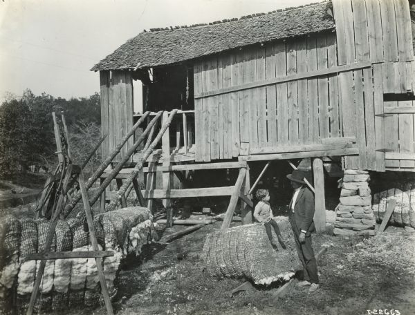A young boy seated on a bale of cotton in front of a farm building looks toward a man standing next to him.