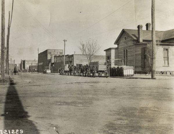 Horse-drawn wagons and bales of cotton line up in the street along the curb near the Lesser-Goldman Cotton Co. Building.