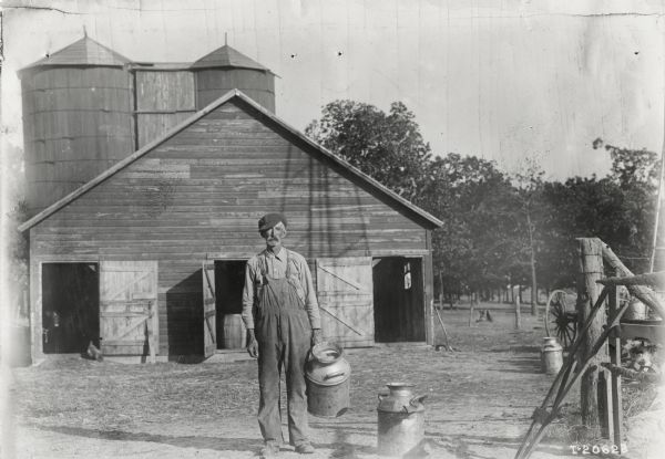 A farmer stands in a barnyard holding a milk can.