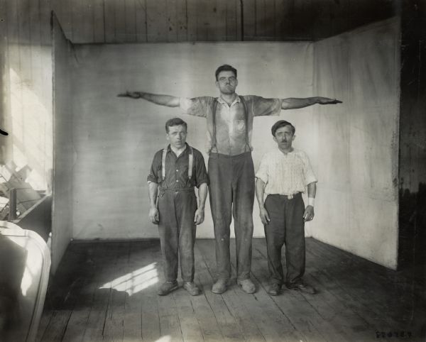 Factory workers from International Harvester's Milwaukee Works demonstrate the difference in their respective heights. The two shorter men, Schultz (left) and Rappel (right) stand under the outstretched arms of the much taller Szalisski. Schultz and Rappel hauled engines around the factory grounds. Szalisski worked in the foundry.