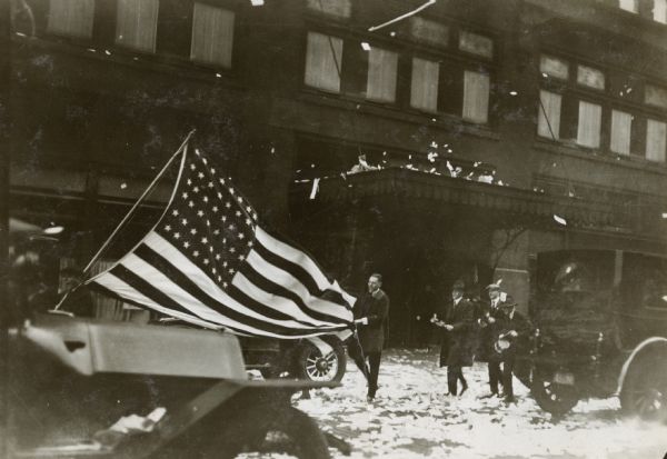 As confetti falls from above, men are walking on Michigan Avenue in a parade. The man on the left is carrying a large American flag on a pole over his shoulder, and another man is holding the back of the flag.
