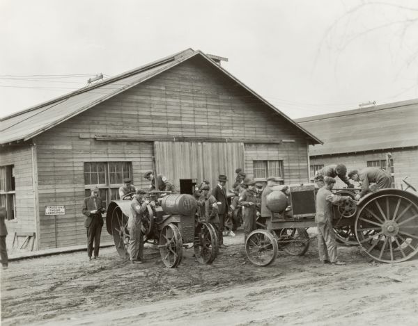 Three groups of male students working on three different tractors outdoors, including a Titan 10-20, while two men (professors?) look on at the Kansas State Agricultural College. Sign on left side of building reads: "Traction Engine Laboratory".