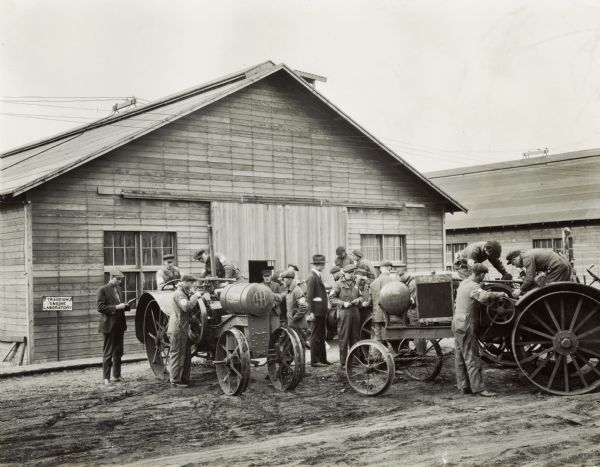 Students working on tractors outdoors, including the Titan 10-20, while two men (professors?) look on at the Kansas State Agricultural College Traction Engine Laboratory. Sign on left side of building reads: "Traction Engine Laboratory".
