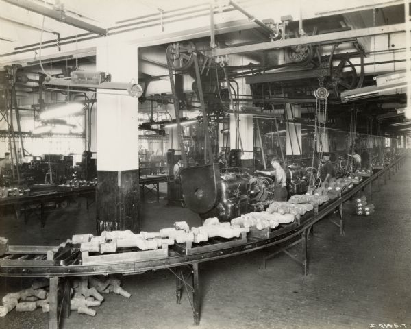 Factory workers stand at work stations while a conveyor moves trays filled with parts.