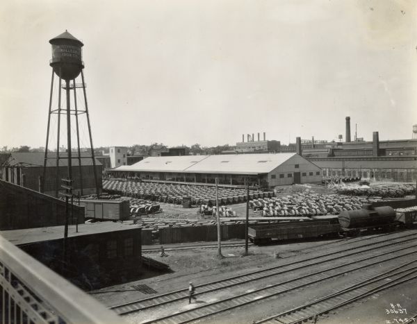 Elevated view of rows of finished tractors cover a yard outside Milwaukee Works. On the left is a water tower, and in the foreground are railroad tracks with rail cars.