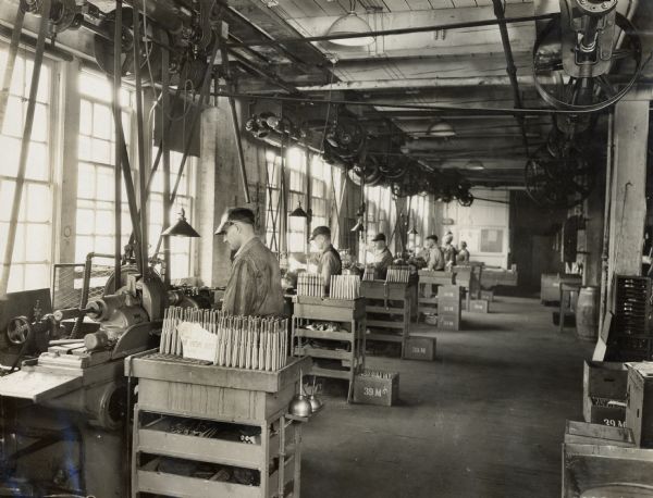 Factory workers standing at work stations at Milwaukee Works. Large windows are along the left wall.