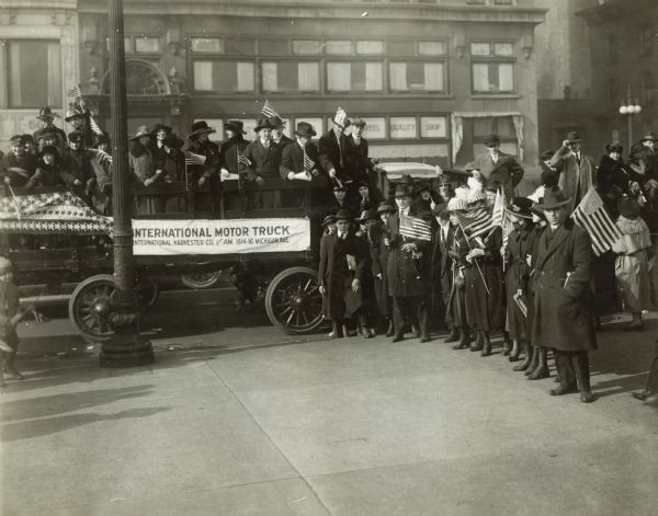 Group of men and women, many holding American flags, on top of and around a wagon or an International truck on a city street. The truck has a banner which reads: "International Motor Truck." The group likely consists of International Harvester employees, and they may have been assembled for a parade on Michigan Avenue, near the company's headquarters building.